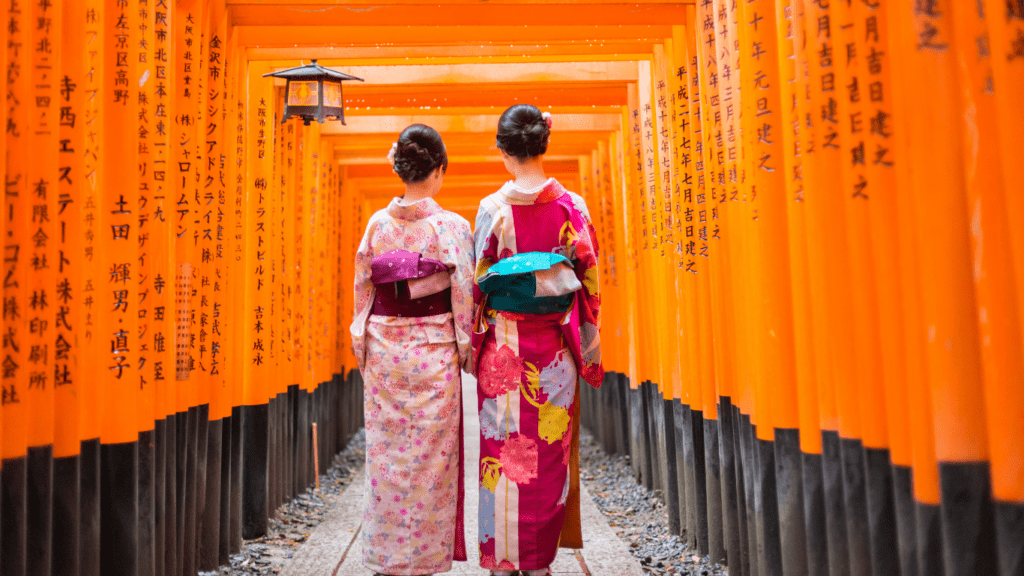 Dos mujeres japonesas de espaldas, vestidas con kimonos tradicionales, caminando entre los emblemáticos torii rojos del santuario Fushimi Inari en Kioto, Japón.