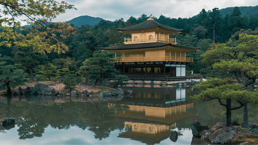 "Templo japonés dorado junto a un río, rodeado de árboles verdes, bajo un cielo claro"