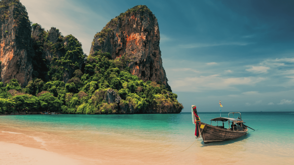 Playa en Tailandia con agua cristalina, una barca tradicional en primer plano, y montañas con bosque al fondo.