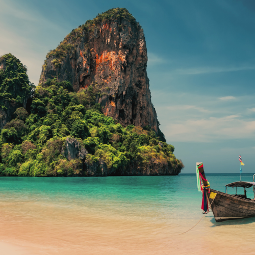 Playa en Tailandia con agua cristalina, una barca tradicional en primer plano, y montañas con bosque al fondo.